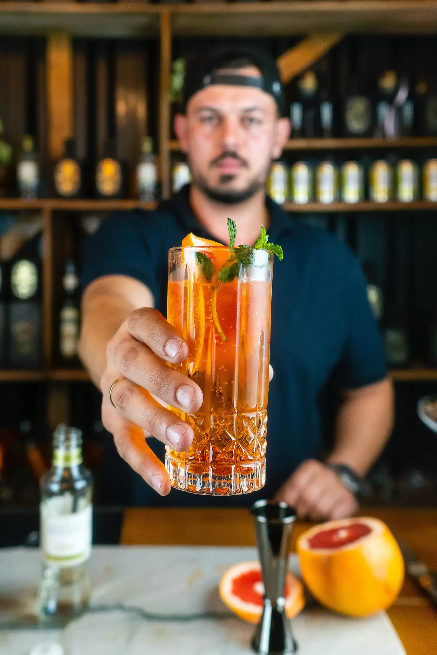 Un barman con cappellino nero offre un cocktail allungato con arancia e menta. Sullo sfondo, uno scaffale pieno di bottiglie in un ambiente accogliente di bar.