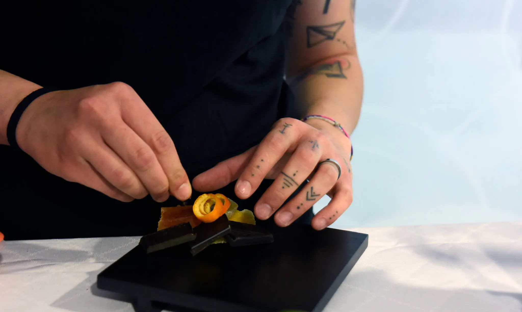 Un bartender con braccia tatuate compone attentamente la guarnizione per un cocktail colorato su un piatto di ardesia.