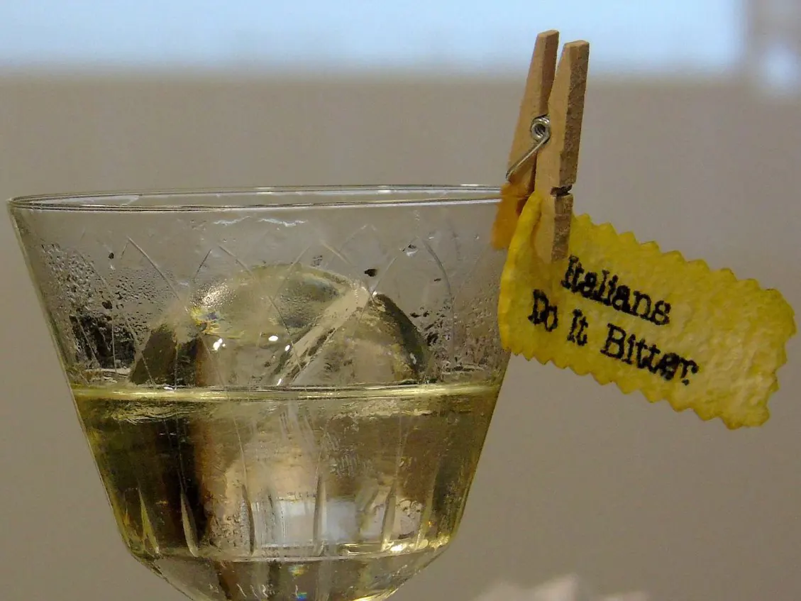 Dettaglio di un bicchiere di cocktail con una guarnizione speciale, con una clip di buccia di limone e la scritta "Italians Do It Bitter".