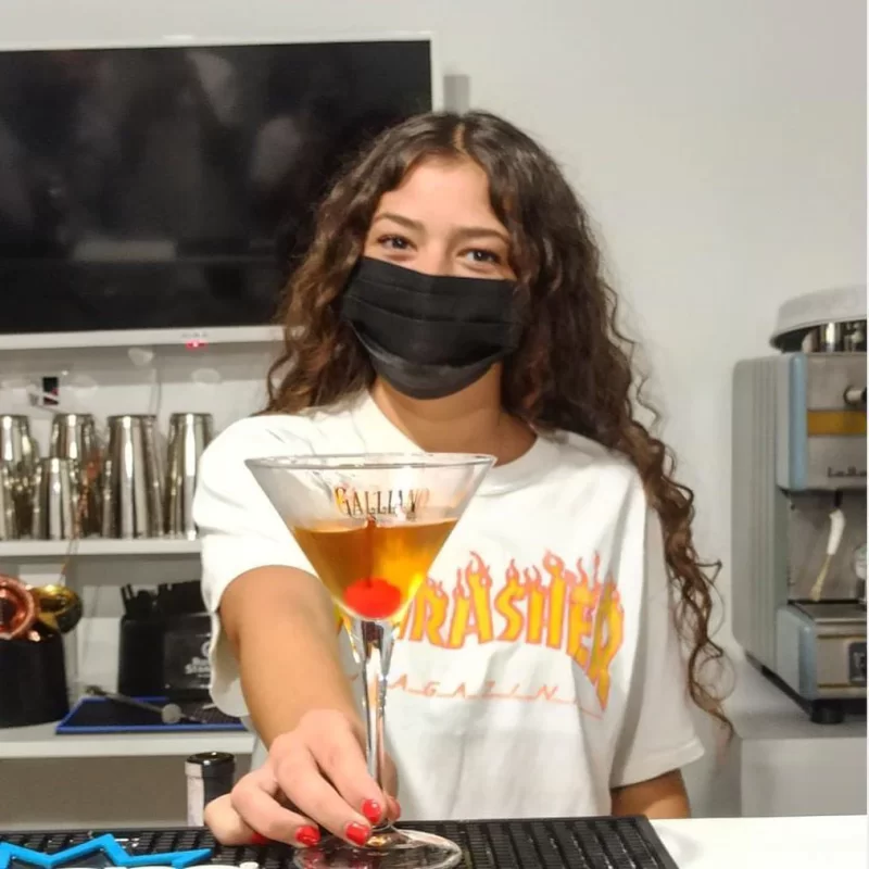 Una giovane studentessa di un corso di barman serve con entusiasmo un cocktail martini con una ciliegina. Il contesto è un'aula didattica con attrezzature da bar professionali sullo sfondo.