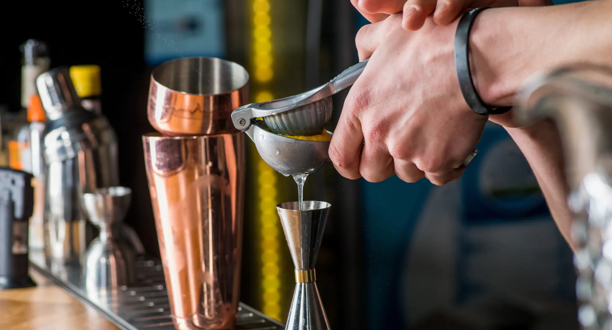 Dettaglio delle mani di un bartender che usa una pressa per agrumi su uno shaker da cocktail in un setup di bar professionale.