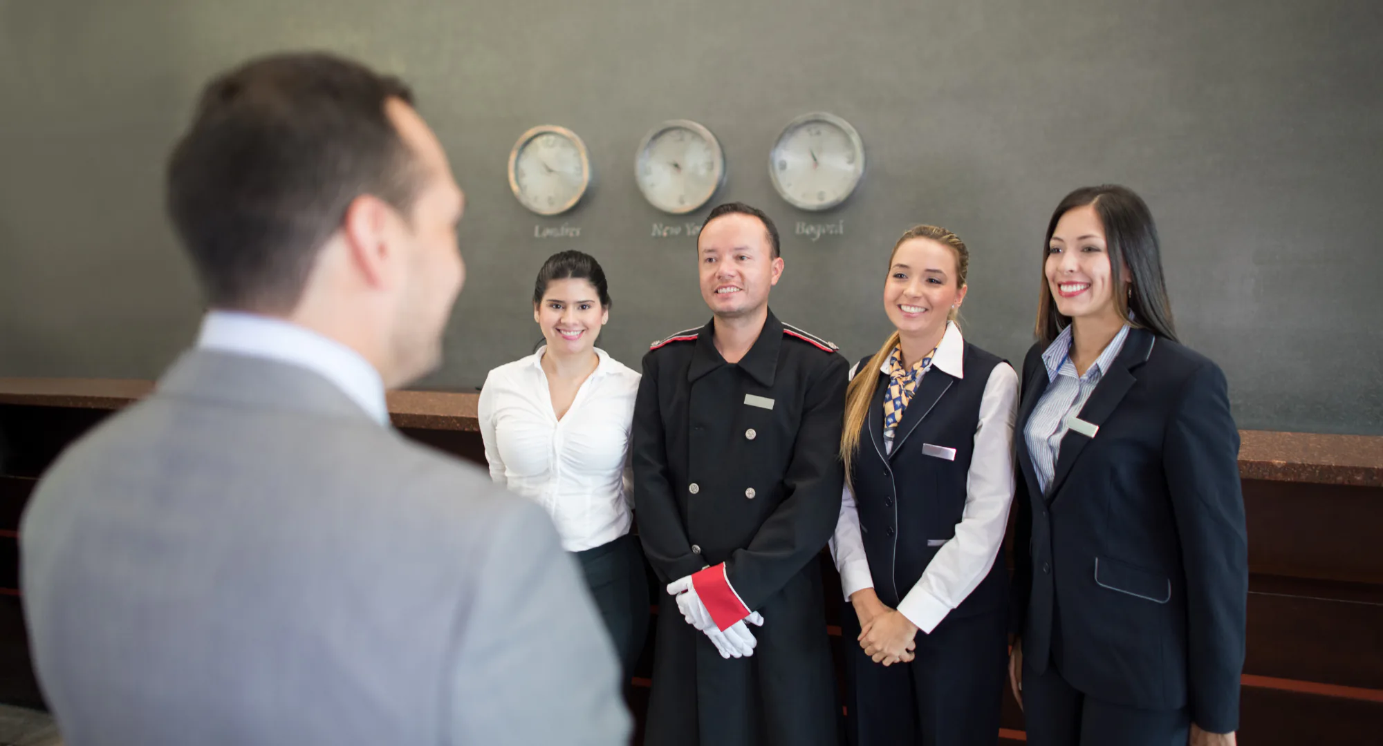 Un team di staff di hotel sorride e saluta il manager alla reception, sotto orologi che mostrano orari internazionali.