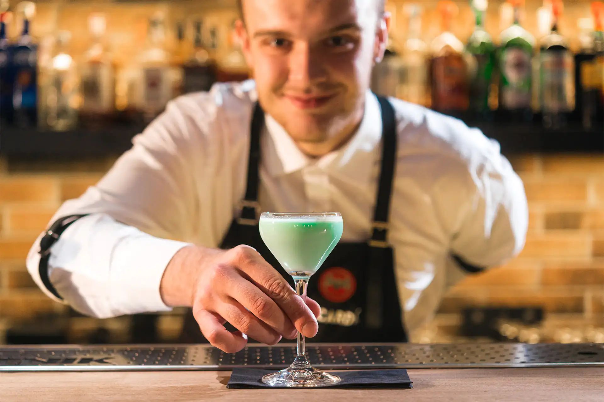 Barman sorridente serve un cocktail verde chiaro in un bicchiere da cocktail in un bar ben fornito.