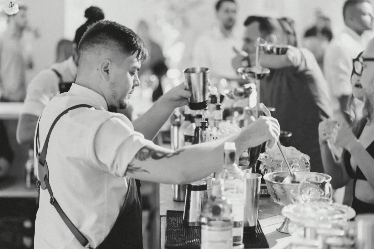 Barman tatuato prepara cocktail in bianco e nero durante un evento affollato, mostrando intensa concentrazione.