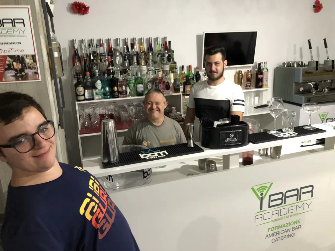 Tre persone di diverse età con sindrome di down sorridenti dietro un bancone di un bar didattico con logo ibaracademy, impegnati nella formazione come barman.