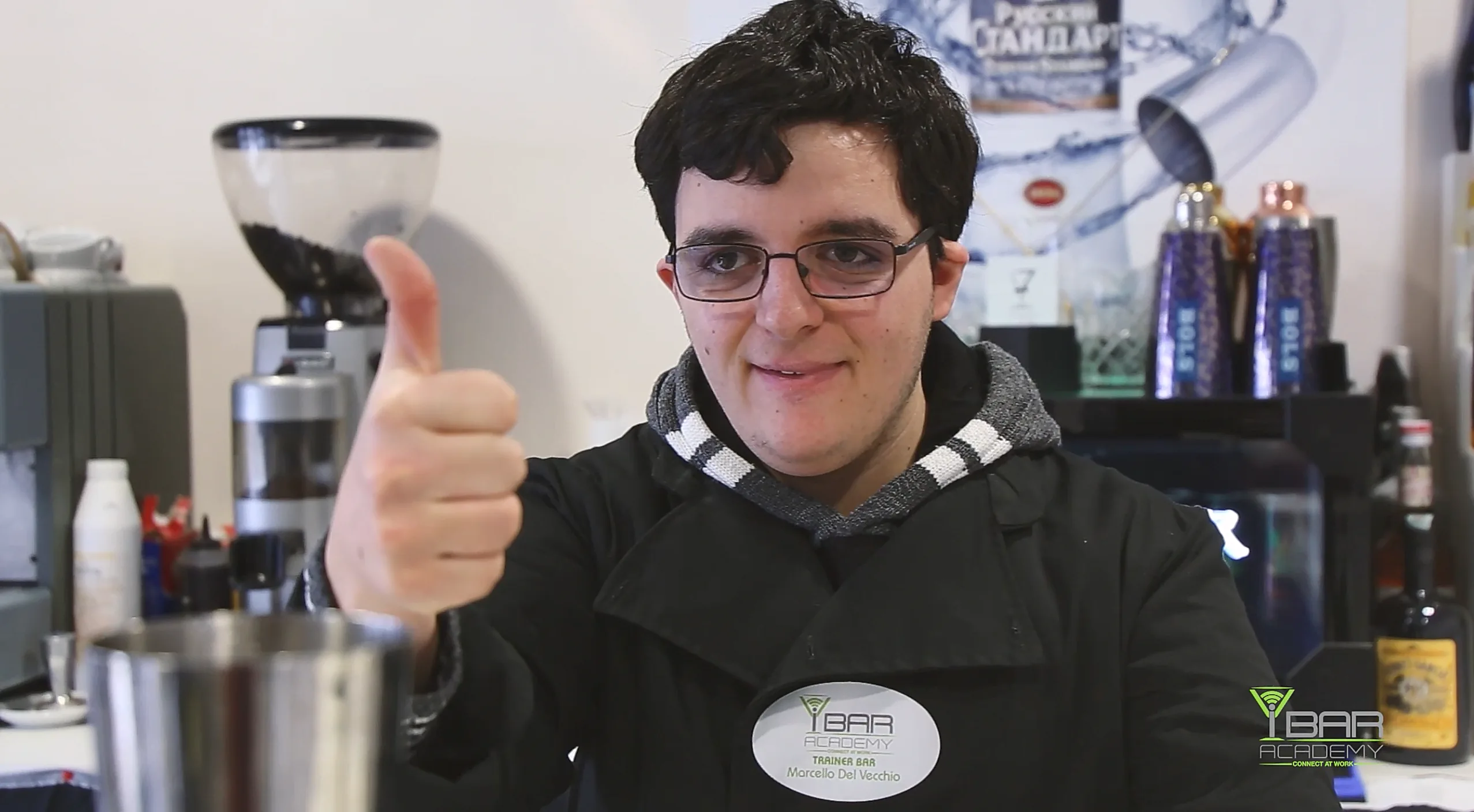 Giovane barman con sindrome di down in uniforme ibaracademy, con un pollice in su, sorridente, davanti a una macchina da caffè in una scuola di baristi.