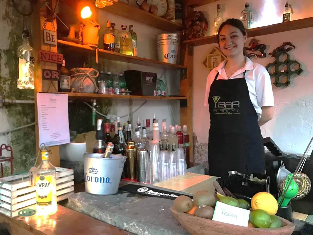 Barista sorridente in grembiule ibaracademy in un bar dal tema rustico, con bottiglie e decorazioni vintage sullo sfondo.