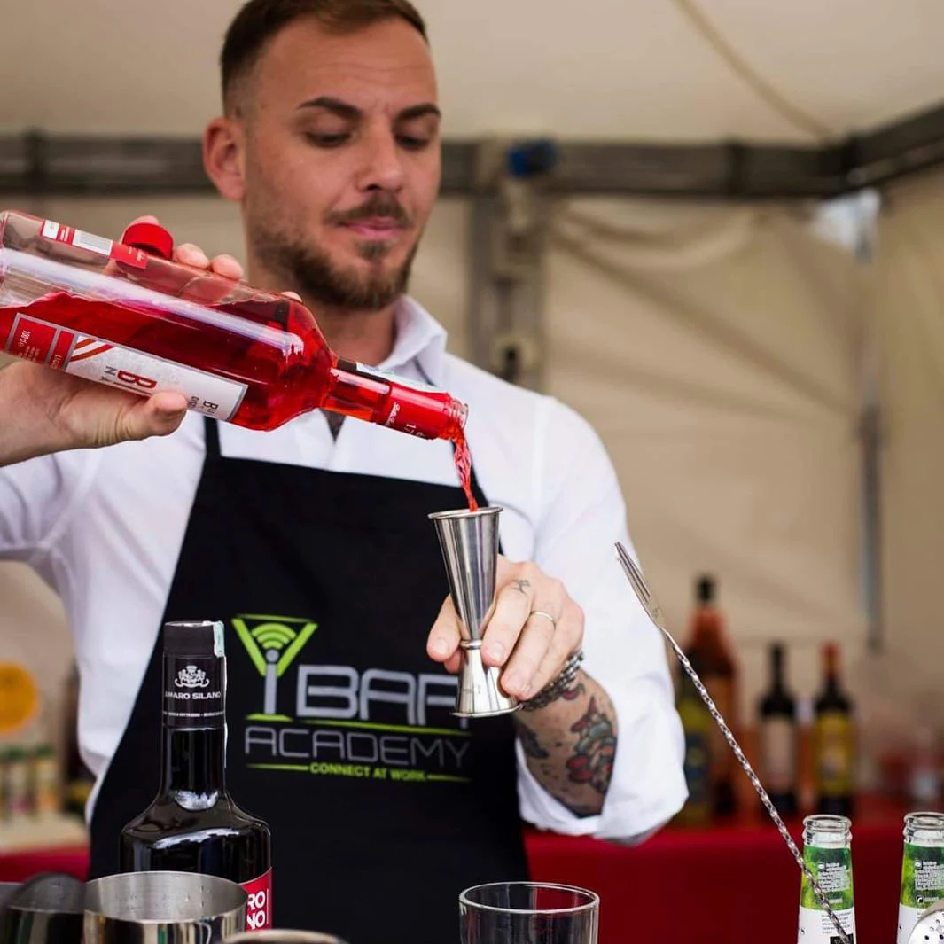 Un formatore professionista di barman, versa un liquido rosso da una bottiglia trasparente in un misurino durante una lezione di mixologia. Indossa un grembiule nero con il logo dell'IBar Academy, simbolo della sua professionalità e del contesto educativo.
